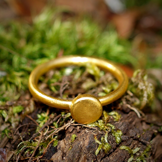 A 24k pure gold band ring with a small round disc design on the top, placed on a mossy surface.